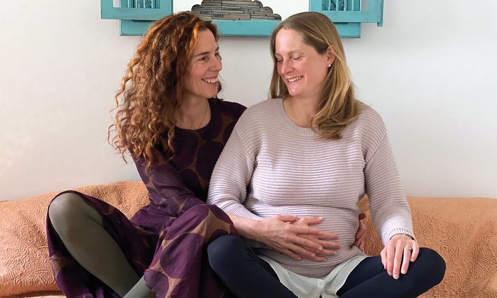 Zwei Frauen sitzen im Schneidersitz nebeneinander, die eine ist schwanger und hält ihre Hand auf ihren Bauch, die andere Frau hält auch ihre Hand auf den Bauch und lächtelt die Schwangere an
