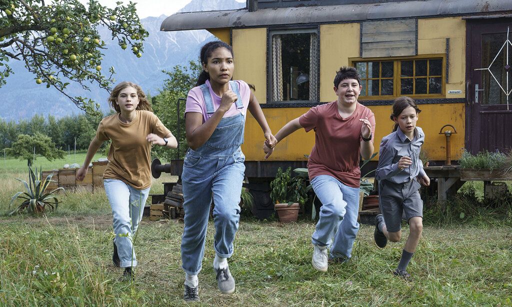 Vier Kinder rennen auf einer Wiese, im Hintergrund ein Wohnwagen und Berge