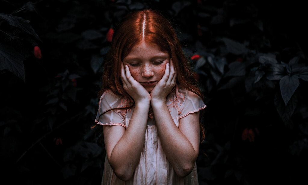 ein Mädchen steht vor einem sehr dunklem Hintergrund, es hat die Augen geschlossen und hält sich die Hände an die Wangen, im hintergrund erkennt man Blätter einer Pflanze