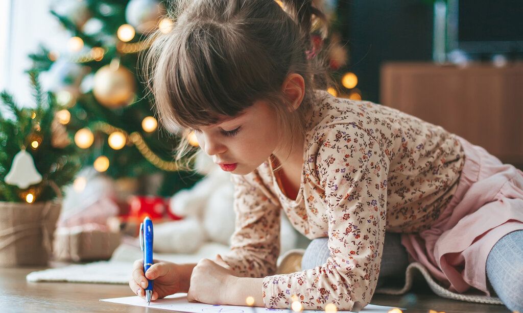 ein kleines Mädchen sitzt auf dem Boden und schreibt einen Brief, im Hintergrund: ein Weihnachtsbaum