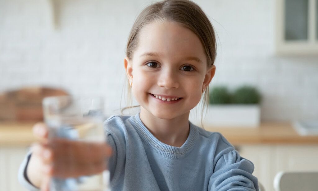 kleines Mädchen lächelt und hält ein Wasserglas in die Kamera