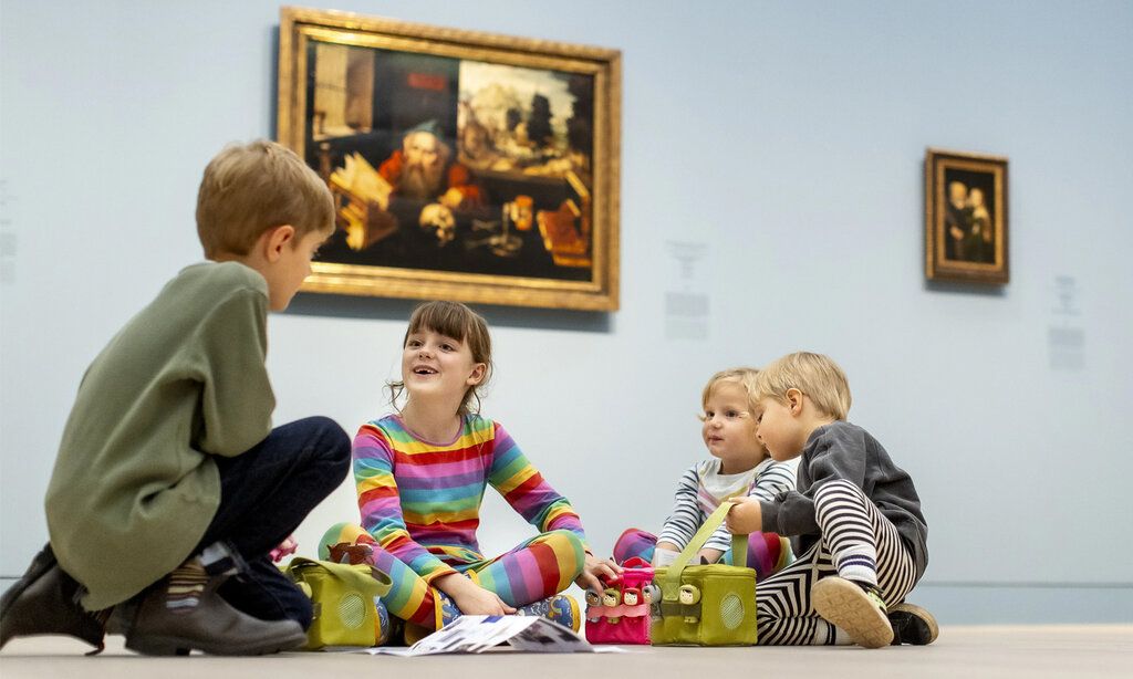 vier Kinder sitzen mit Audioguides auf dem Boden im Kunstpalast, im Hintergrund Bilder alter Meister