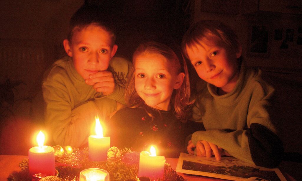 drei Kinder sitzen zusammen, vor ihnen brennende Kerzen vom Adventskranz, dunkle gemütliche Atmosphäre