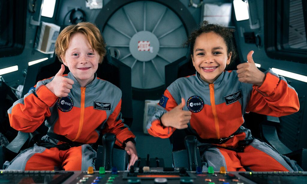 Junge und Mädchen sitzen mit Astronautenanzügen im Cockpit eines Raumschiffe und halten die Daumen hoch