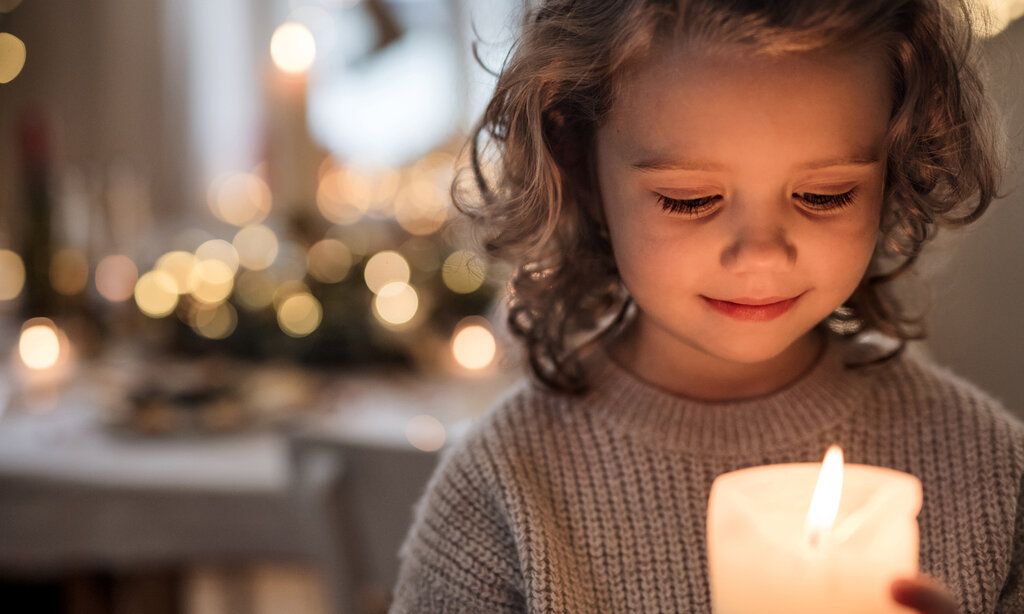 ein Mädchen hält eine Kerze vor sich und schaut sie an, im Hintergund weihnachtliche Lichter