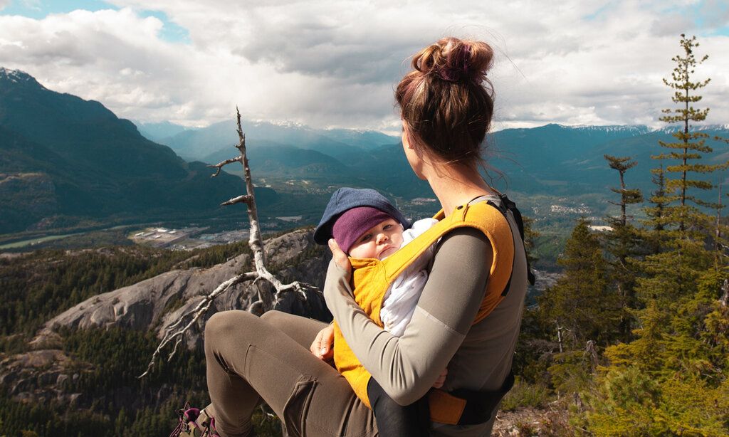 Frau mit Baby in Babytrage sitzt auf einem Berg und guckt in die Landschaft, das Baby guckt in die Kamera