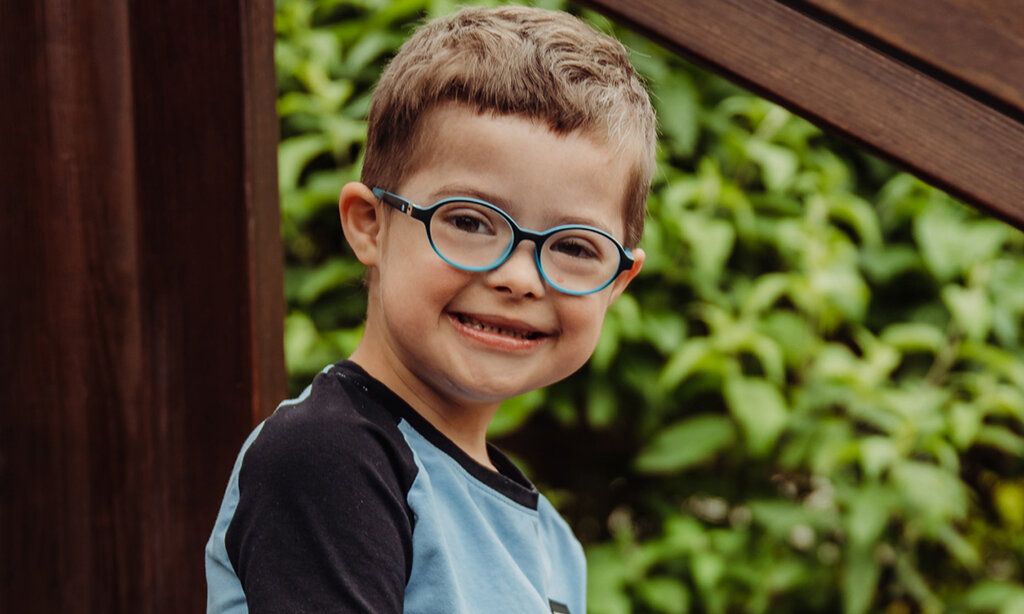 Porträt eines Jungen mit Down-Syndrom, er trägt eine Brille und lächelt in die Kamera, im Hintergrund Gebüsch