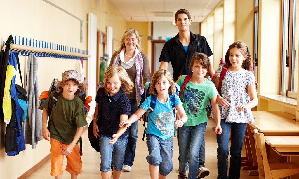 fünf Kinder rennen durch den Flur einer Schule, der Kamera entgegen, hinter ihnen gehen eine Lehrerin und ein Lehrer