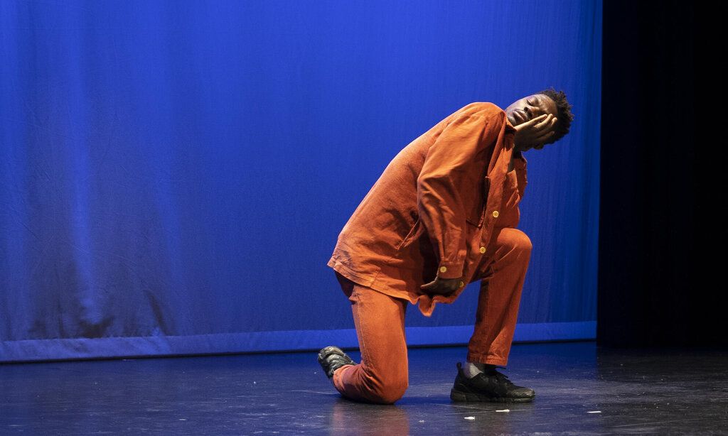 Ein Mann in orangefarbener Kleidung kniet mit geschlossenen Augen auf der Bühne.