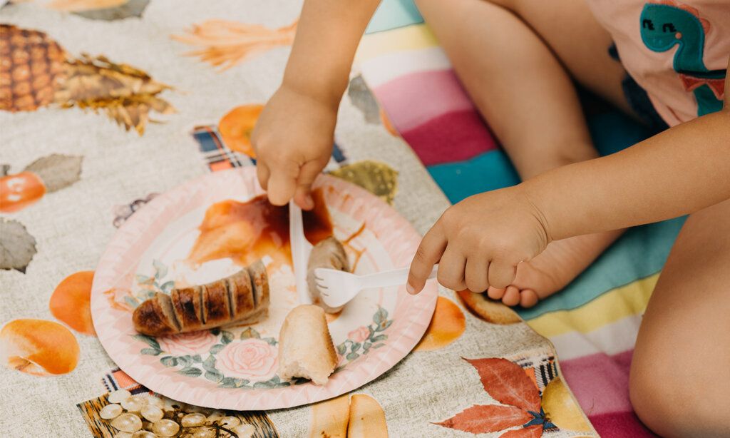 Detailaufahme, ein Kind sitz auf einer Picknickdecke und schneidet eine Bratwurst
