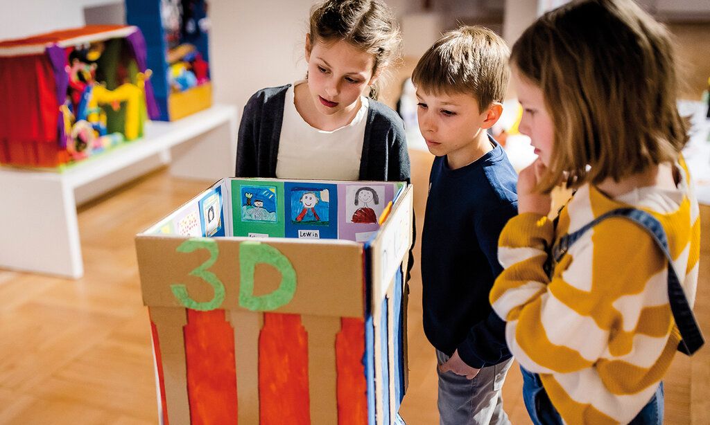 Drei Kinder stehen im Museum und gucken sich ein Kunstwerk aus Pappe von einem Kind an.