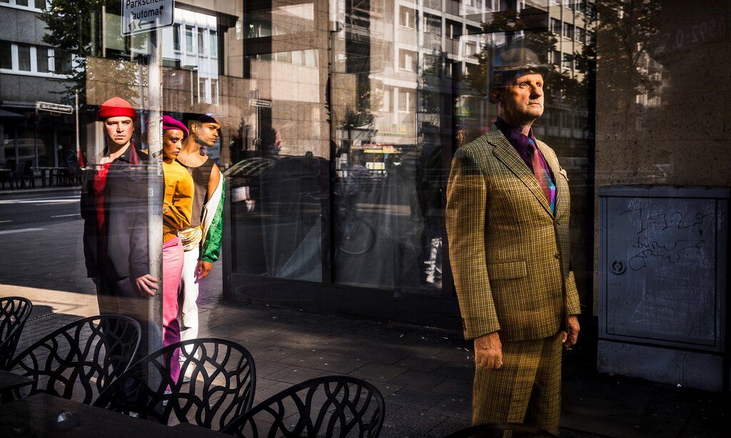 Ein Mann mit Melone und kariertem Anzug steht vor einem Schaufenster, hinter seinem Rücken blicken drei Kinder auf ihn.