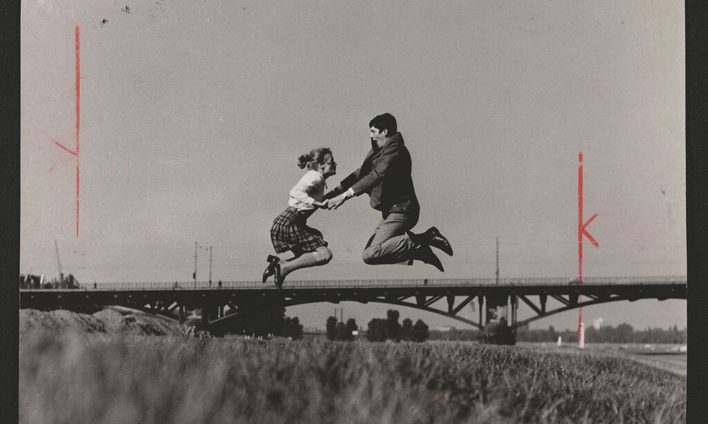 Ein schwarz-weiß Bild zeigt eine Frau und einen Mann die sich an den Händen haltend ansehen und auf einer Wiese vor einer Brücke in die Luft springen