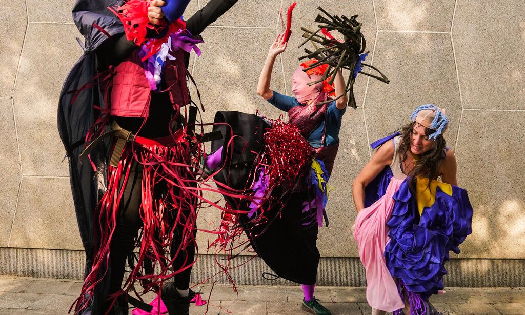 Drei Tänzerinnen und Tänzer bewegen sich vor einer Steinmauer, eingehüllt in verschiedene Stoffe