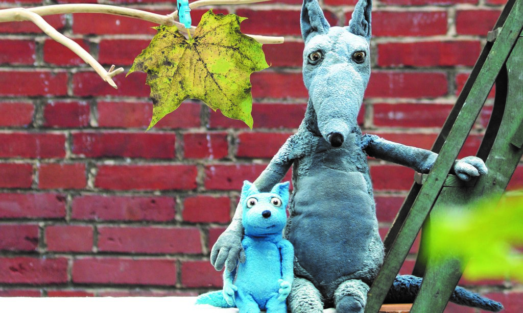 Der große Wolf und der kleine Wolf sitzen Arm in Arm vor einer Backsteinmauer.