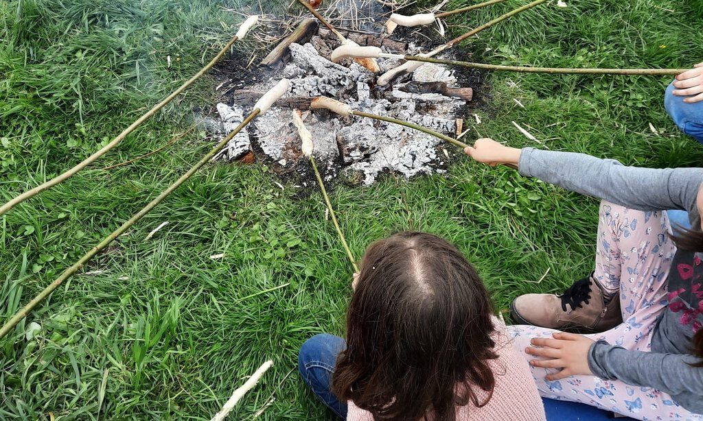 Kinder sitzen an einem Lagerfeuer und grillen Würstchen am Stock