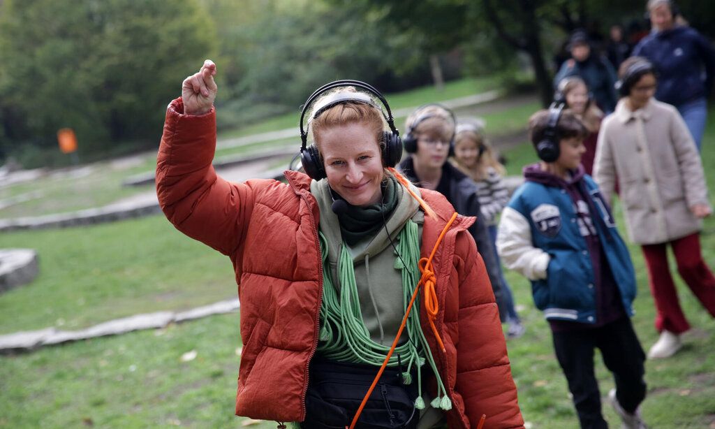 Eine Frau mit vielen Kabeln um den Hals und Kopfhörern auf den Ohren läuft lächelnd vor einer Gruppe Kinder über eine Wiese und hebt dabei einen Arm.