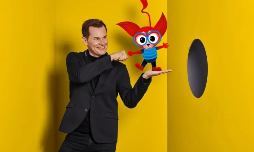 Malte Arkona steht vor einer gelben Wand mit der Kunstfigur Mezzo auf seiner Hand.