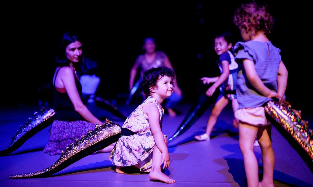 Kleine Kinder bewegen sich mit auf den Rücken geschnallten Reptilienschwänzen über die Bühne.