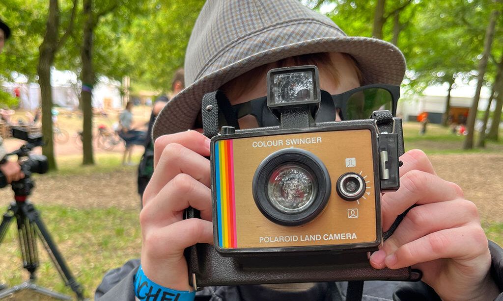 Ein als Detektiv verkleideter Junge fotografiert mit einer Polaroid Kamera im Park.