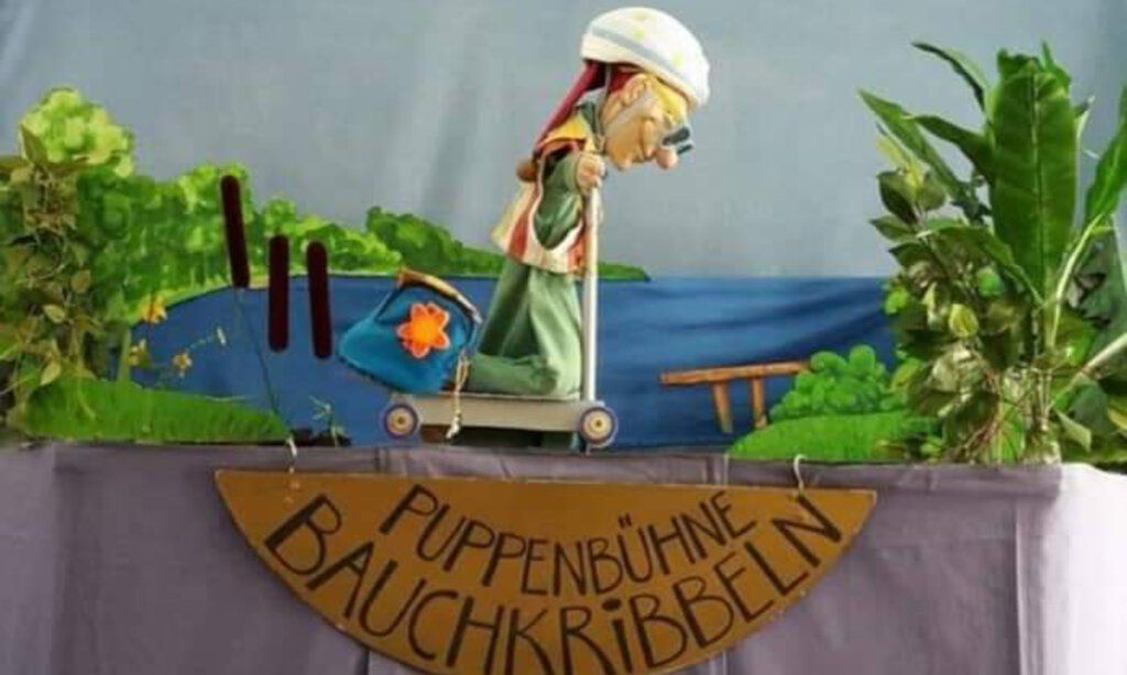 Bühnenkulisse der Puppenbühne Bauchkribbeln mit Kasperle auf einem Roller mit Helm und Sonnenbrille.