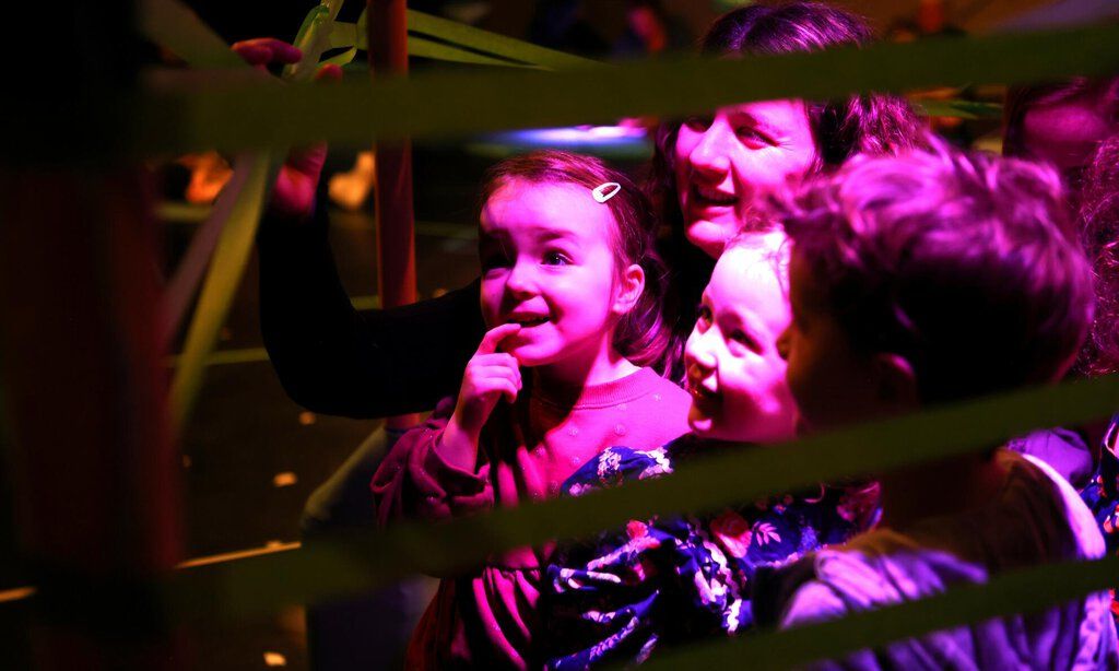 Drei kleine Kinder stehen mit ihrer Mutter staunend in einem Gewirr von Klebestreifen, die sich durch den Raum ziehen