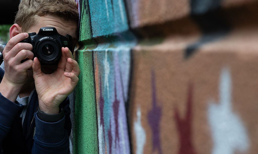 Ein junger Mann fotografiert Graffiti an einer Hauswand lehnend.