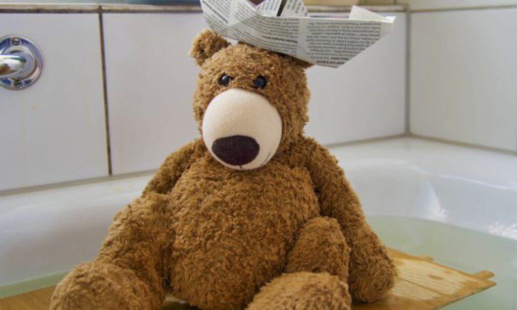 Ein Teddybär mit Papierhut sitzt auf einem Stück Holz in einer vollen Badewanne