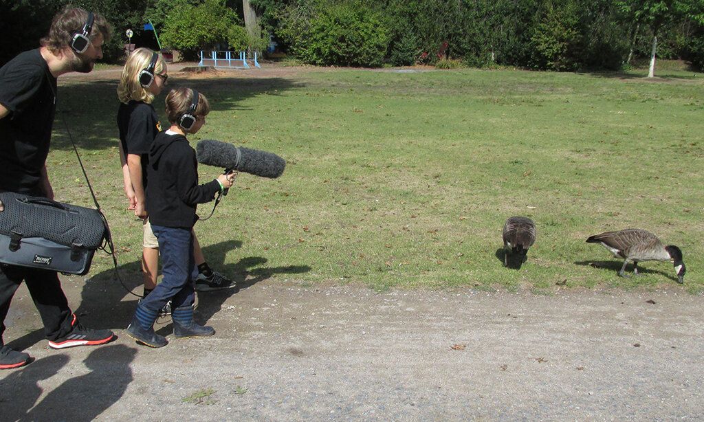Zwei Kinder und ein Tontechniker nehmen mit Mikrofon und Headsets die Geräusche zweier Gänse in einem Park auf.