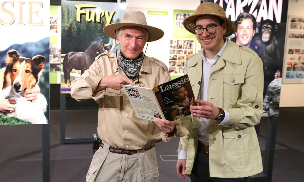 Zwei Männer stehen  in Safari-Anzügen im Filmmuseum vor alten Filmplakaten, in der Hand ein Buch über Lassie