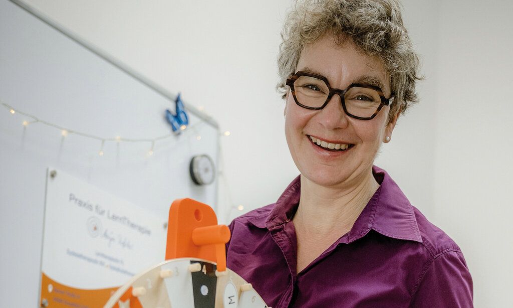 Frau mit auffälliger Brille lacht in die Kamera, im Hintergrund ein White Board