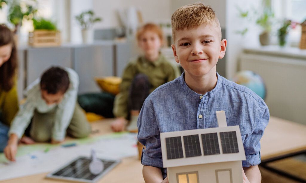 Ein Junge hält ein Modell von einem Haus mit Solarenergie-Panelen in den Händen, im Hintergrund weitere Kinder