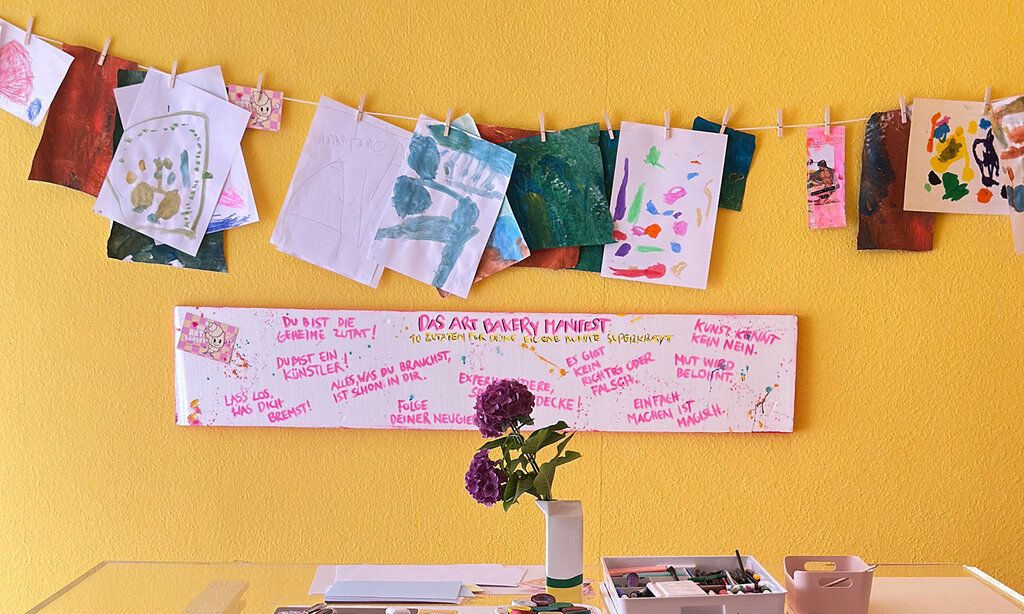 Art Bakery, Tisch mit Malutensilien und Hortensie vor gelber Wand, gespannte Leine mit Kinderbildern