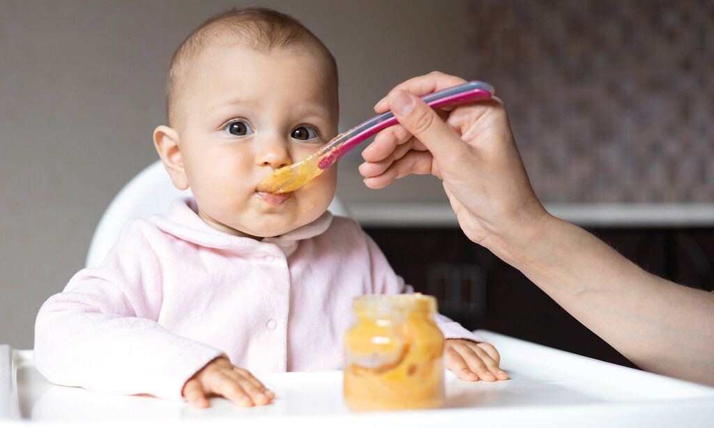 Baby wird mit Brei gefüttert und guckt dabei in die Kamera, der Arm einer erwachsenen Person führt den Löffeln zum Mund des Babys