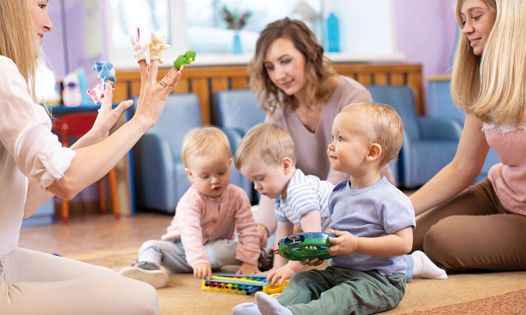 Drei Betreuerinnen und drei Kleinkinder sitzen auf dem Boden spielen zusammen, eine Betreuerin hat Fingerpuppen auf den Fingern