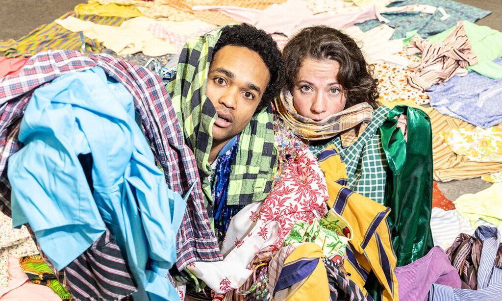 ein Mann und eine Frau liegen auf dem Boden, umgeben und überschüttet von Kleidungsstücken