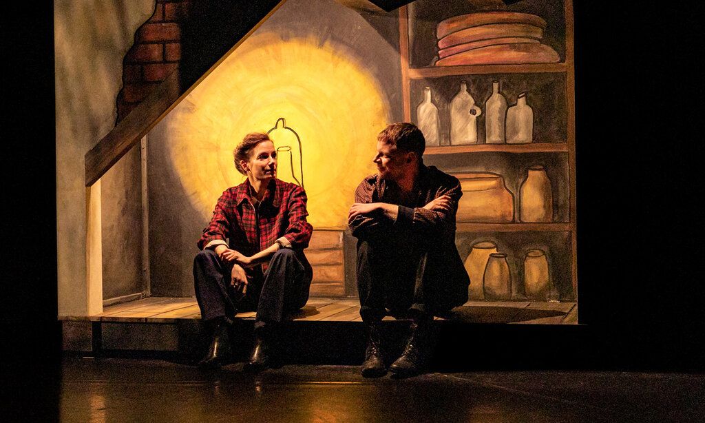 eine Schauspielerin und ein Schauspieler sitzen auf der Bühne, im Hintergrund eine gemalte Kulisse von einer Hütte