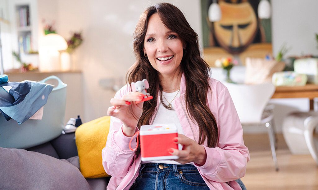 Carolin Kebekus sitzt in einem Wohnzimmer und hält eine Toniebox mit einer kleinen Elefantenfigur in der Hand