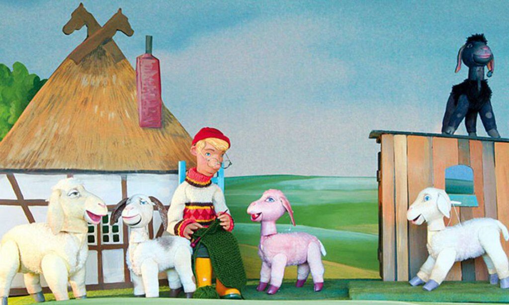 Eine Schäfer-Puppe sitzt strickend vor der Kulisse, umringt von Schafspuppen.