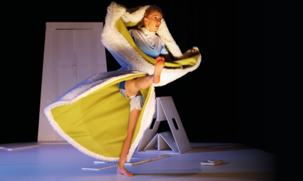 Eine Tänzerin umhüllt von einem gelben Umhang mit Fell tanzt barfuß auf der Bühne