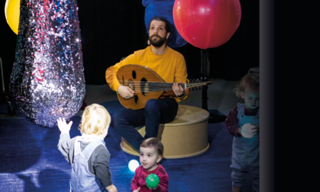 Ein Mann mit Ukulele spielt und singt inmitten von kleinen Kindern und Luftballons