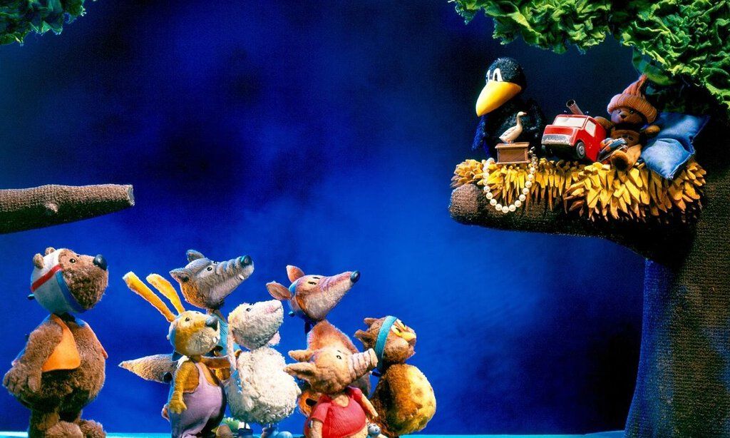 Verschiendene Waldtiere stehen vor einem Baum und starren hinauf auf einen Ast, auf dem Rabe Socke sitzt.