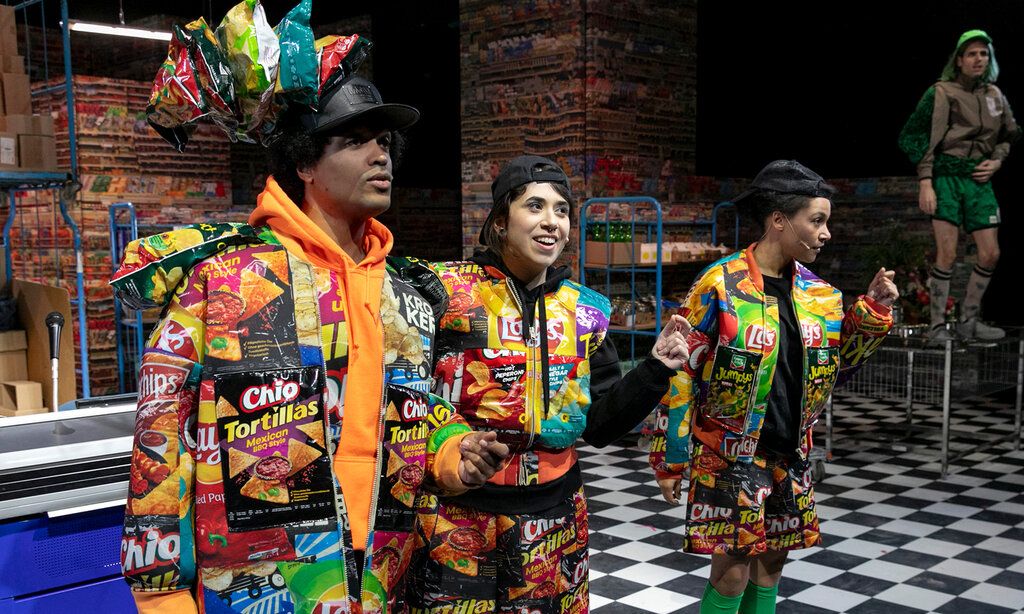 drei Schauspieler:innen auf einer Bühne, die wie ein Supermarkt aussieht, die Kleidung besteht aus Chipstüten