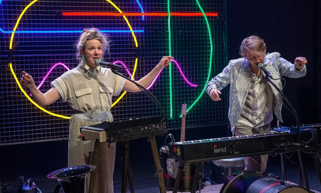 eine Musikerin und ein Musiker machen zusammen Musik, im Hintergund bunter Neonröhren