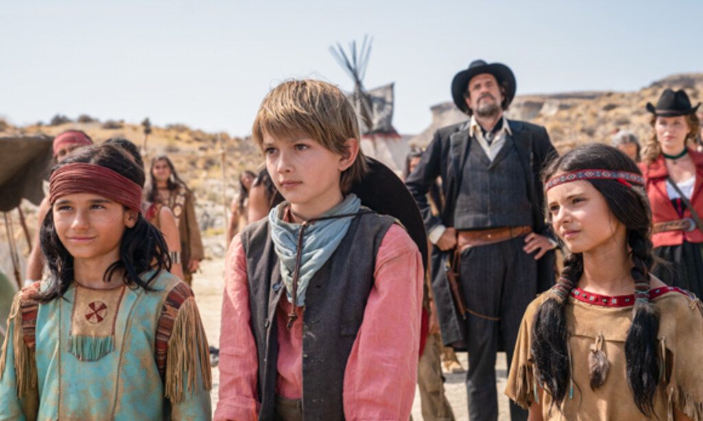 Drei Kinder in Westernkostümen in der Prärie im Hintergrund Erwachsene