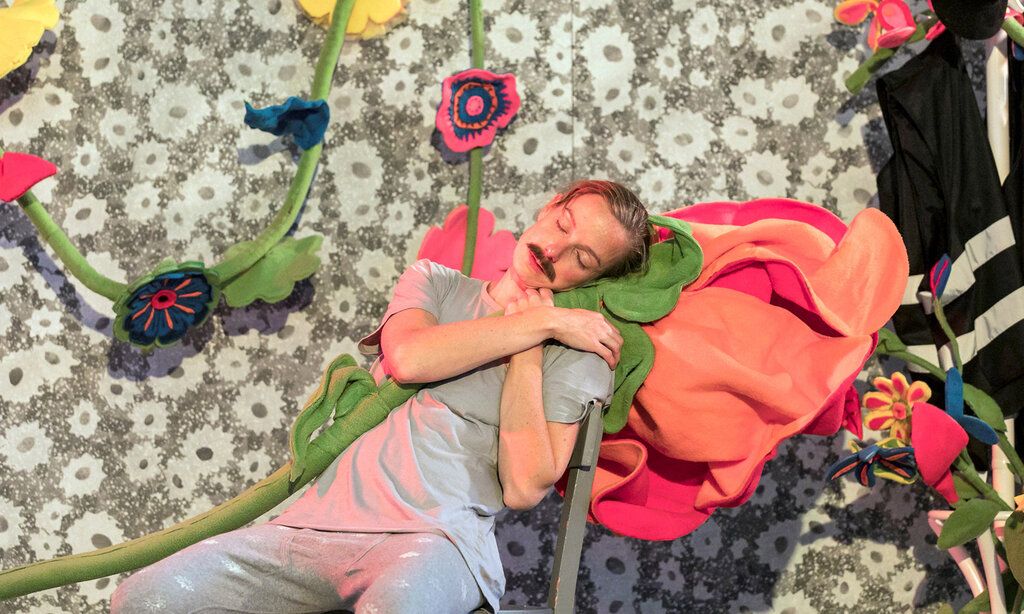 eine Schauspielerin mit einem Schnurrbart schläft auf einem Stuhl und hält dabei eine Stoffblume im Arm, im Hintergrund weitere Stoffblumen