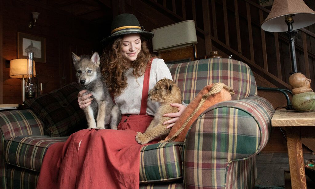 eine junge Frau mit Hut sitzt mit einem jungen Wolf und einem jungen Löwen auf einem karierten Sofa