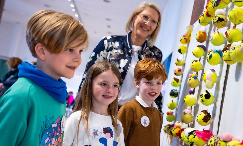 Drei Kinder der Klasse 2b der St. Mauritius Schule und eine Frau stehen vor einem Kunstwerk, das aus Tennsibällen besteht