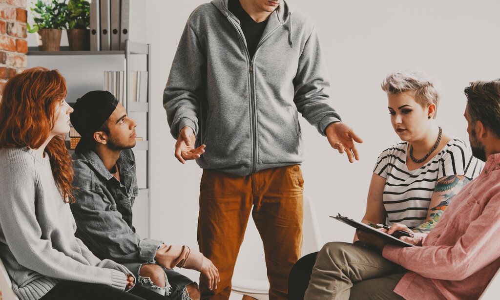 fünf Erwachsene sitzen zusammen im Gespräch, ein Mann in der Mitte steht und redet