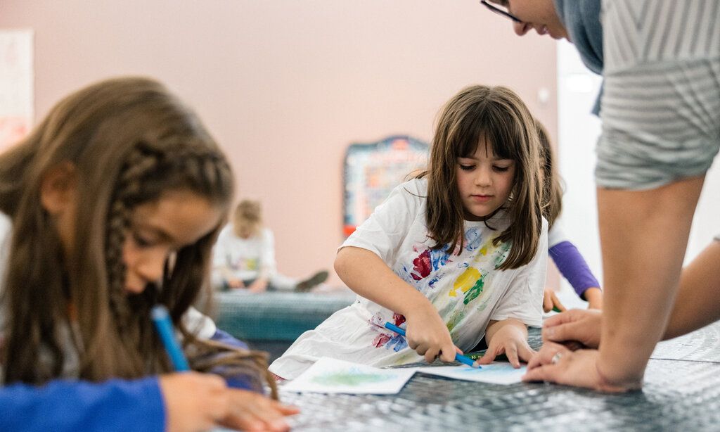 Kinder sitzen zusammen und zeichnen, eine Frau spricht mit einem Mädchen
